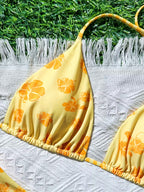 A yellow bikini top with an orange hibiscus floral print, laid flat on a white textured blanket with white fringe, set against a background of green grass.
