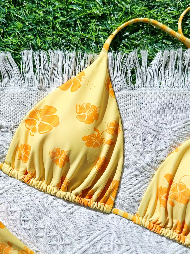 A yellow bikini top with an orange hibiscus floral print, laid flat on a white textured blanket with white fringe, set against a background of green grass.
