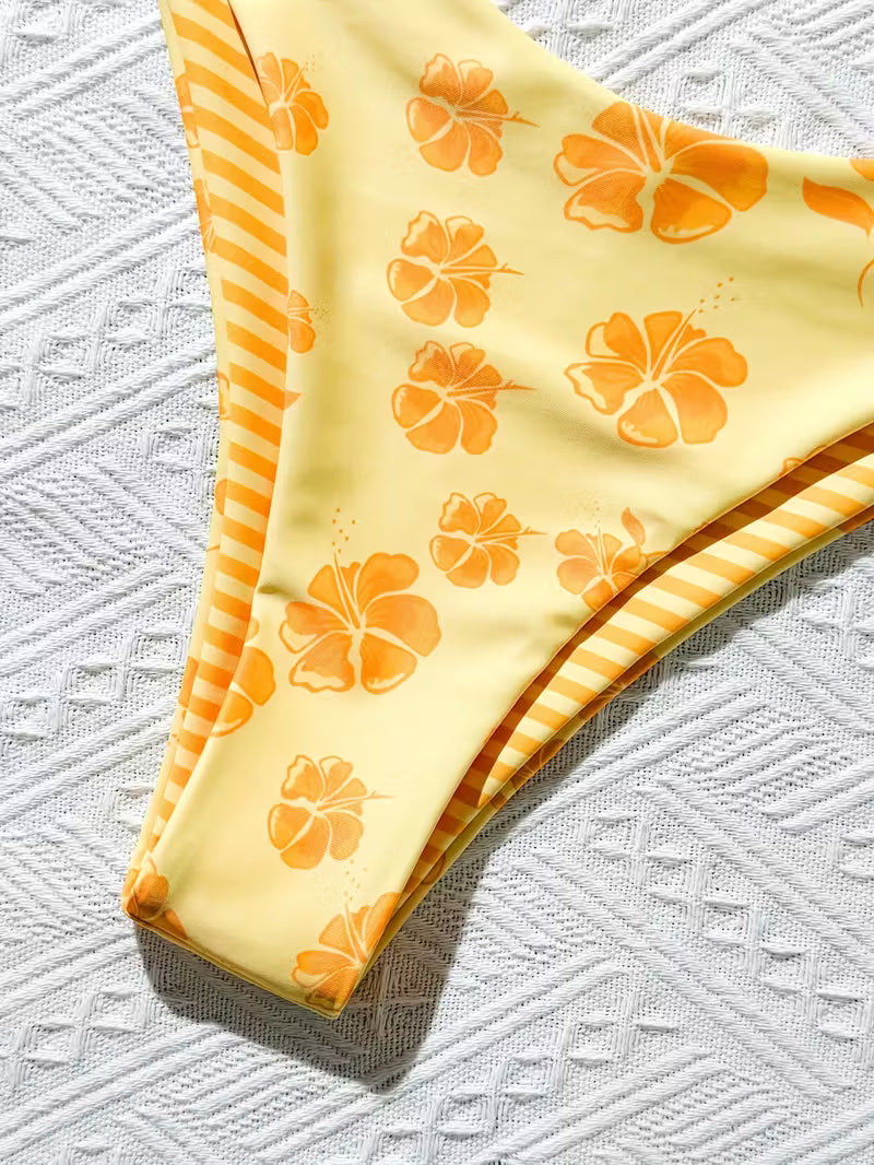 A close-up of a bikini bottom in pale yellow with orange hibiscus floral print and orange-and-yellow striped trim, laid flat on a textured white fabric background.